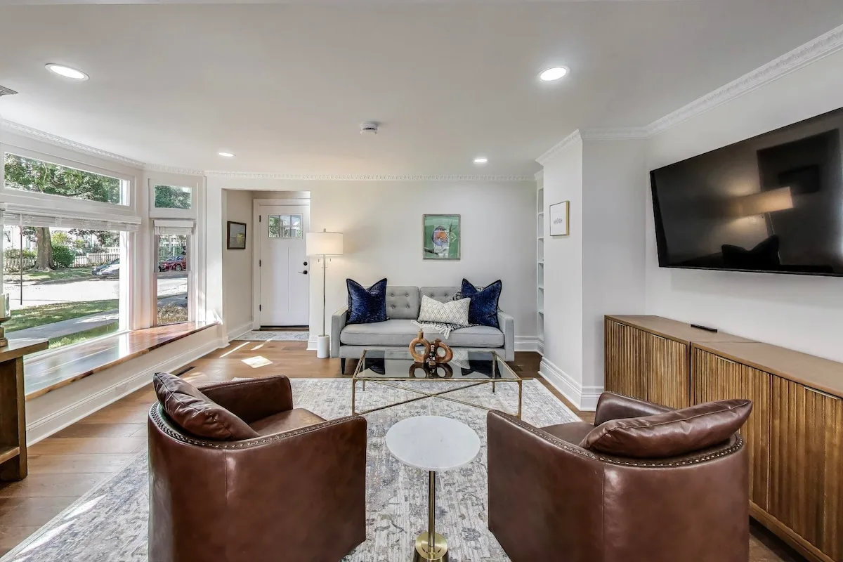 Bright living room with a gray sofa, leather armchairs, and a bay window looking onto a tree-lined street