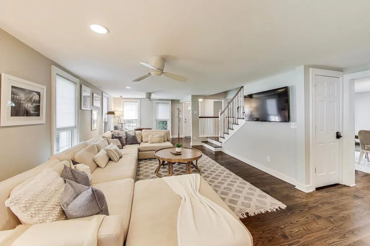 Bright living room with a beige sectional sofa, round wood coffee table, and a staircase with white spindles