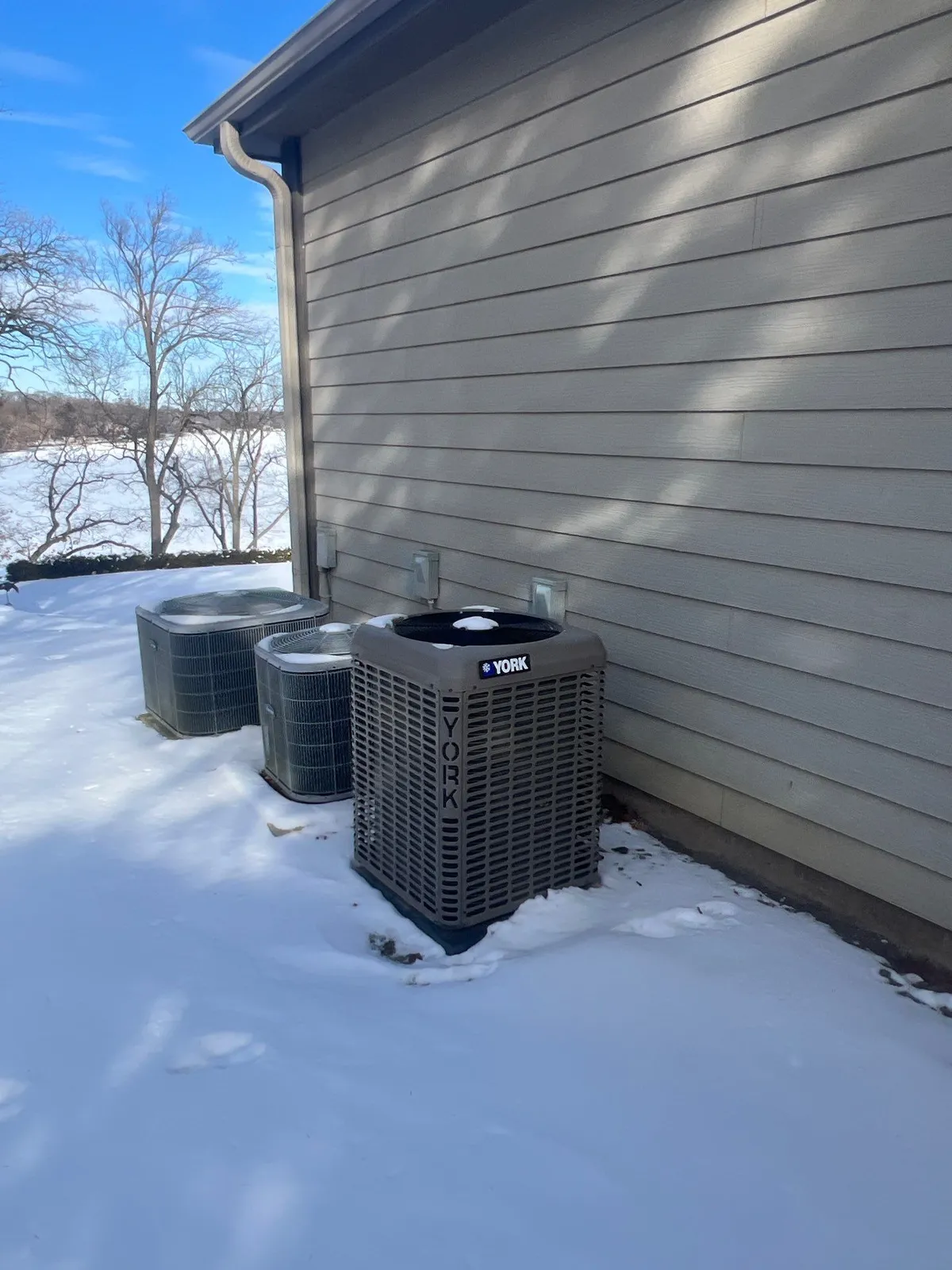 Three outdoor air conditioning condenser units lined up along a snow-covered wall during a winter home watch visit